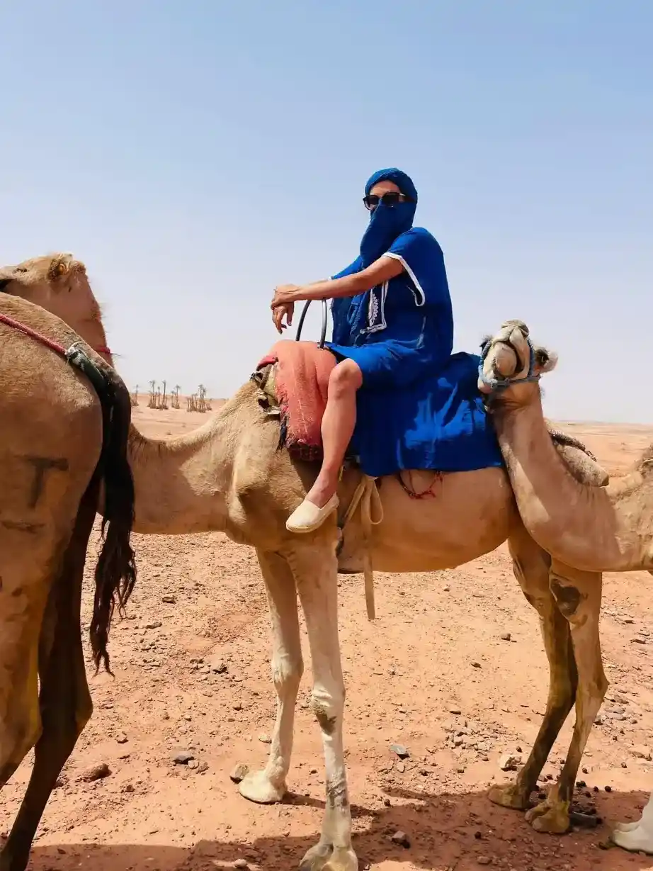 Tourist dressed in a blue tuareg robe riding a camel through the sandy desert landscape of Marrakech's Palmeraie