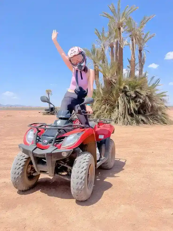 Happy woman in a helmet waving while riding a red quad bike in the Palmeraie of Marrakech on a sunny day