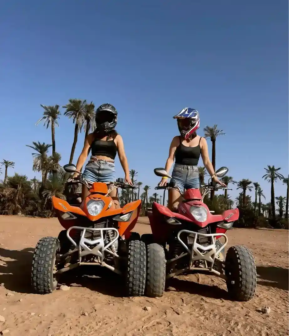 Two women wearing helmets standing on orange and red quad bikes in the desert terrain of Marrakech's Palmeraie